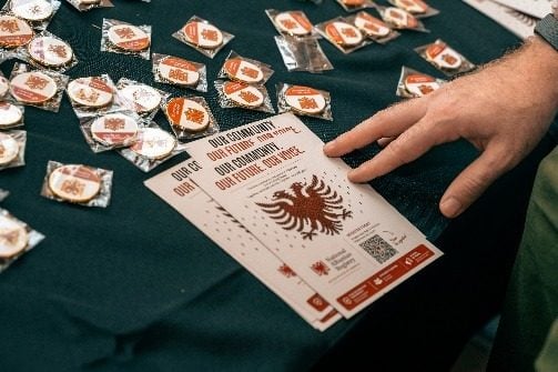Një dorë mbi tavolinën me pinat e Regjistrit Kombëtar Shqiptar dhe një kartë "Count Me Albanian".