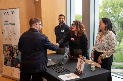 Një përfaqësues i Vialto Partners duke shtrënguar dorën me një pjesëmarrës te tavolina e sponsorit.