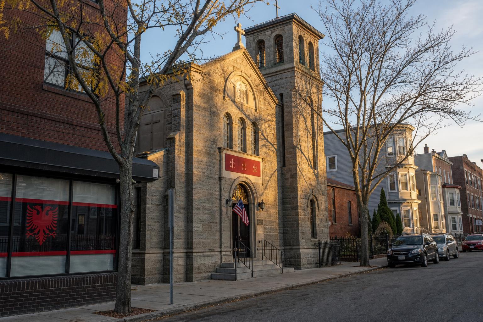 Stone Albanian Orthodox parish church on a quiet South Boston block at golden hour, with a small American flag at the doorframe and an adjacent storefront showing an Albanian double-headed eagle decal in the window.