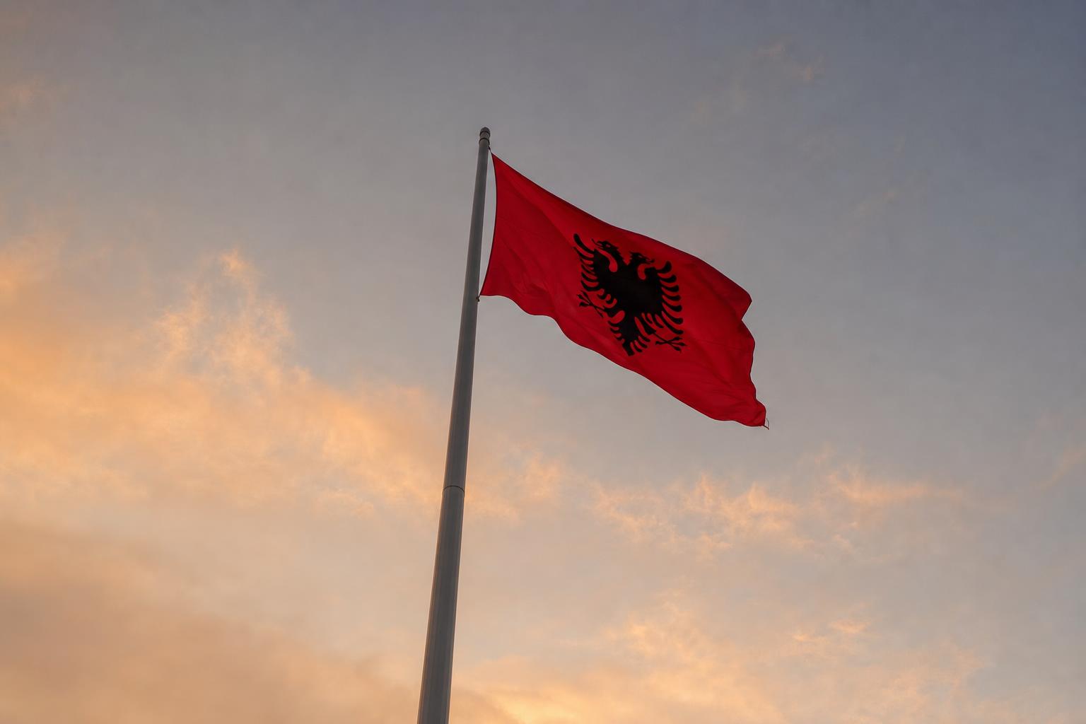 Tall flagpole with the Albanian flag flying gently against a pale dusk sky, view from below looking up.