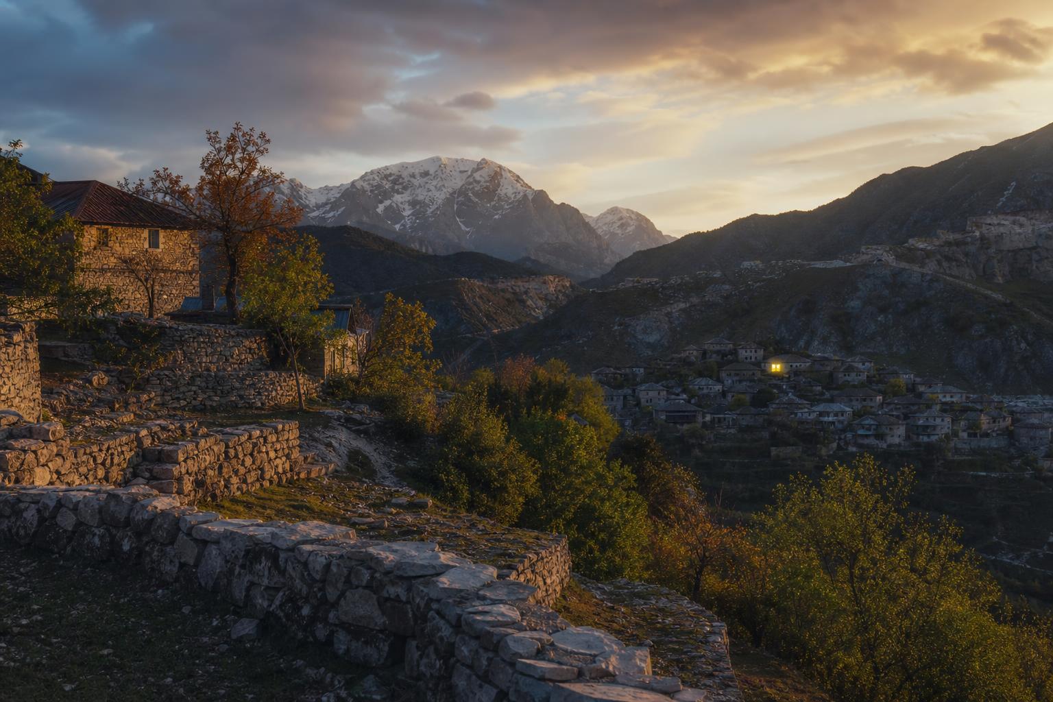 Rugged Albanian alpine landscape at dusk — stone terraces, a distant village with one window glowing, snow-capped peaks in the deep background.
