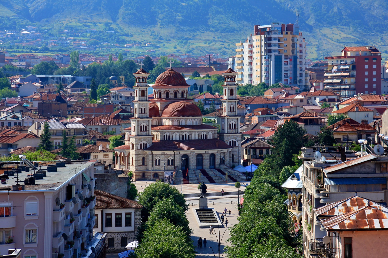 Exterior view of the Cathedral of the Resurrection of Christ in Korçë, Albania — a tall pale-stone Eastern Orthodox church with twin bell towers and a central dome, sited at the head of a long pedestrian boulevard. Korçë is the city Eliza Dushku's paternal grandparents emigrated from to Boston in the early 20th century, part of the same Korçë-to-Massachusetts migration that founded Vatra in 1912.