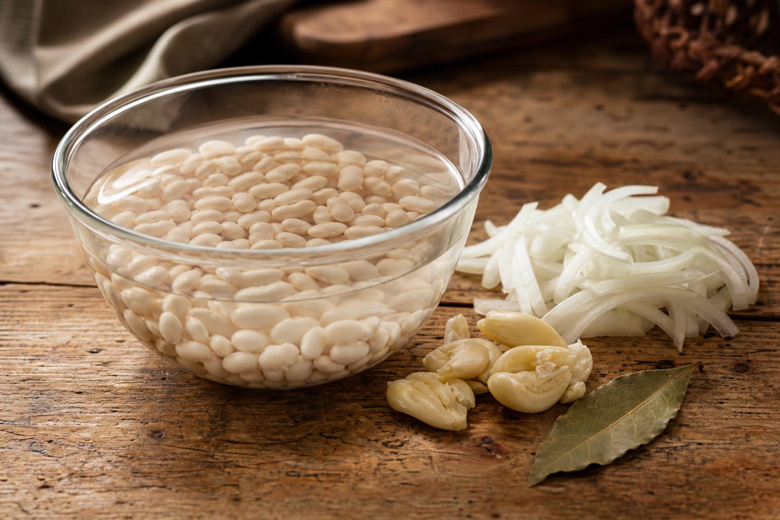 Dried white beans soaking in a clear glass bowl beside a small pile of sliced onions, smashed garlic cloves, and a single bay leaf, on a wooden table.