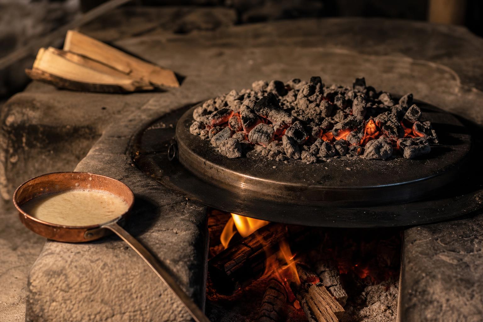Traditional saç (metal lid) over an open wood fire with hot embers piled on top of the lid, a copper ladle of batter beside it on a stone hearth.