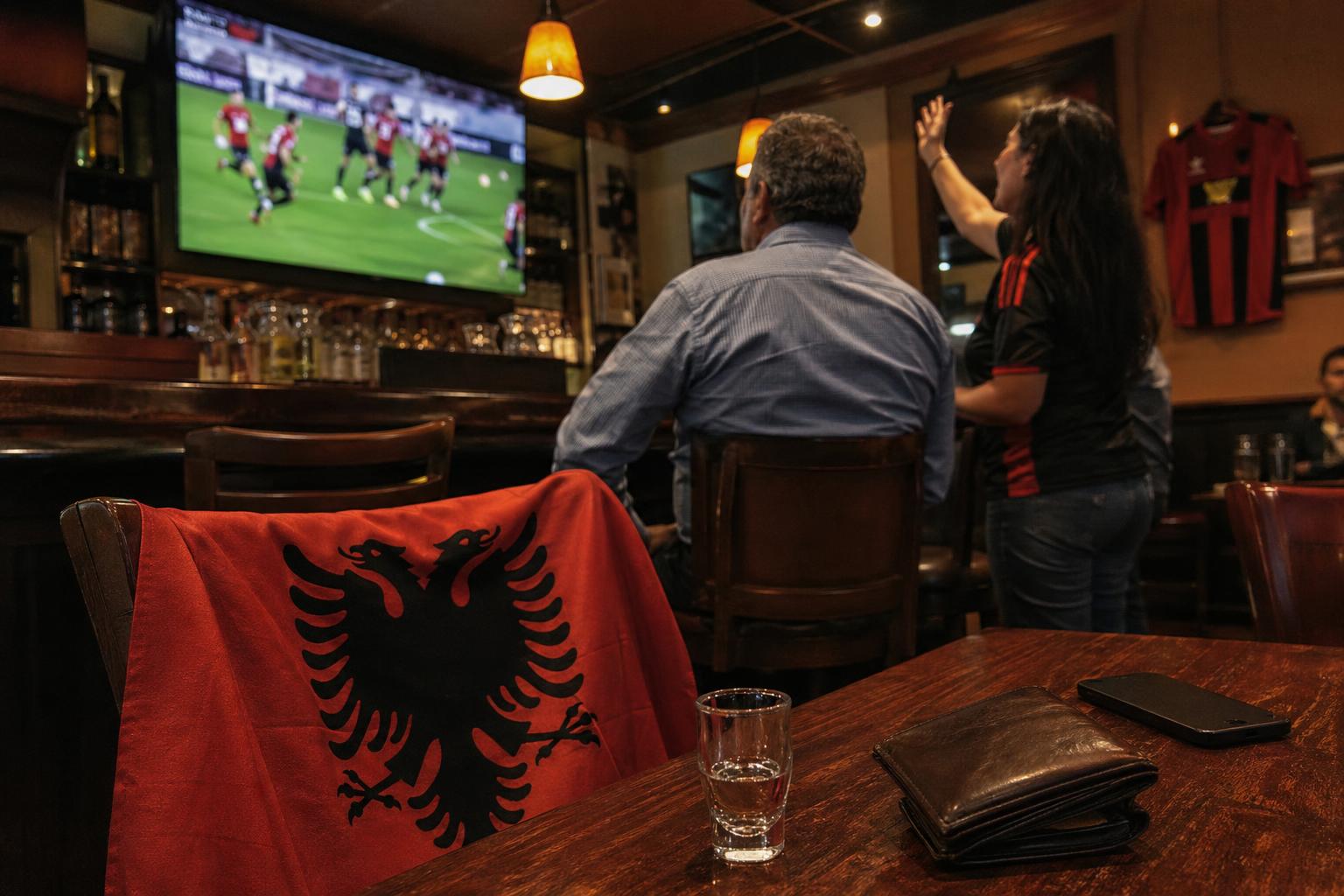 Albanian flag draped over a bar chair with a match playing on the TV behind it
