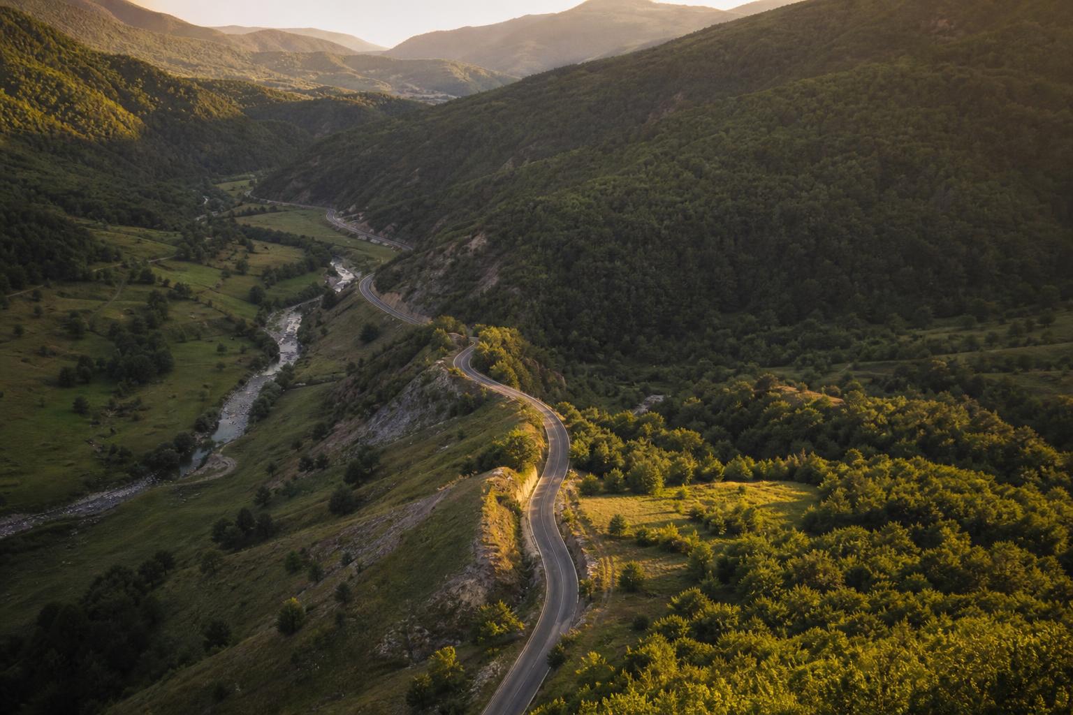 Aerial view of the Albania-Kosovo border landscape at golden hour — two valleys meeting, a winding road between them, wooded hills, no buildings dominant.