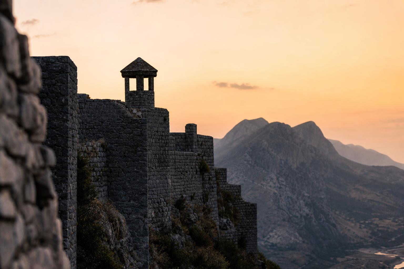 Rugged Albanian mountain stronghold at dusk — silhouette of stone walls and a watchtower against a pale orange sky.